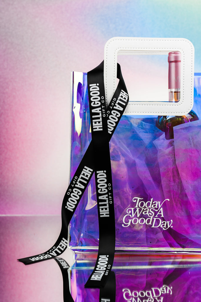 holographic gift bag with Today was a good day written on the front. Wine bottle inside the bag. Black ribbon draped through the handles with white text "Hella Good Gift Co"