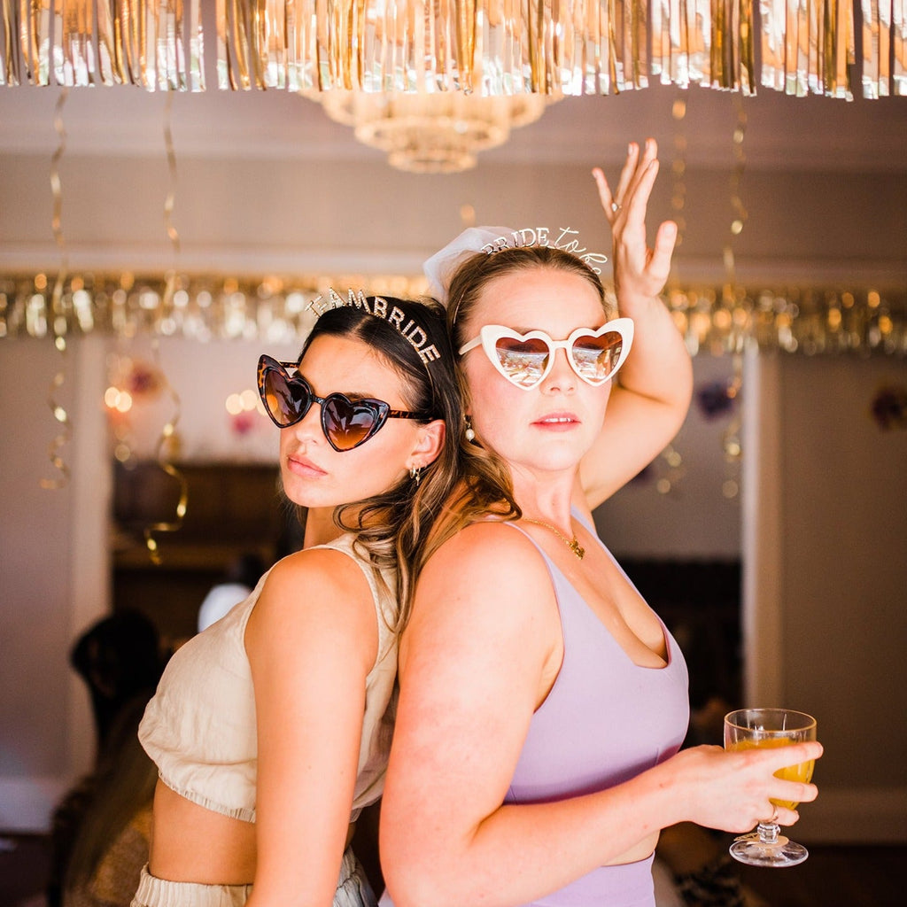 Two cute girls wearing heart-shaped sunglasses and matching 'Team Bride' headbands with gold decorations, representing bridesmaid and bride