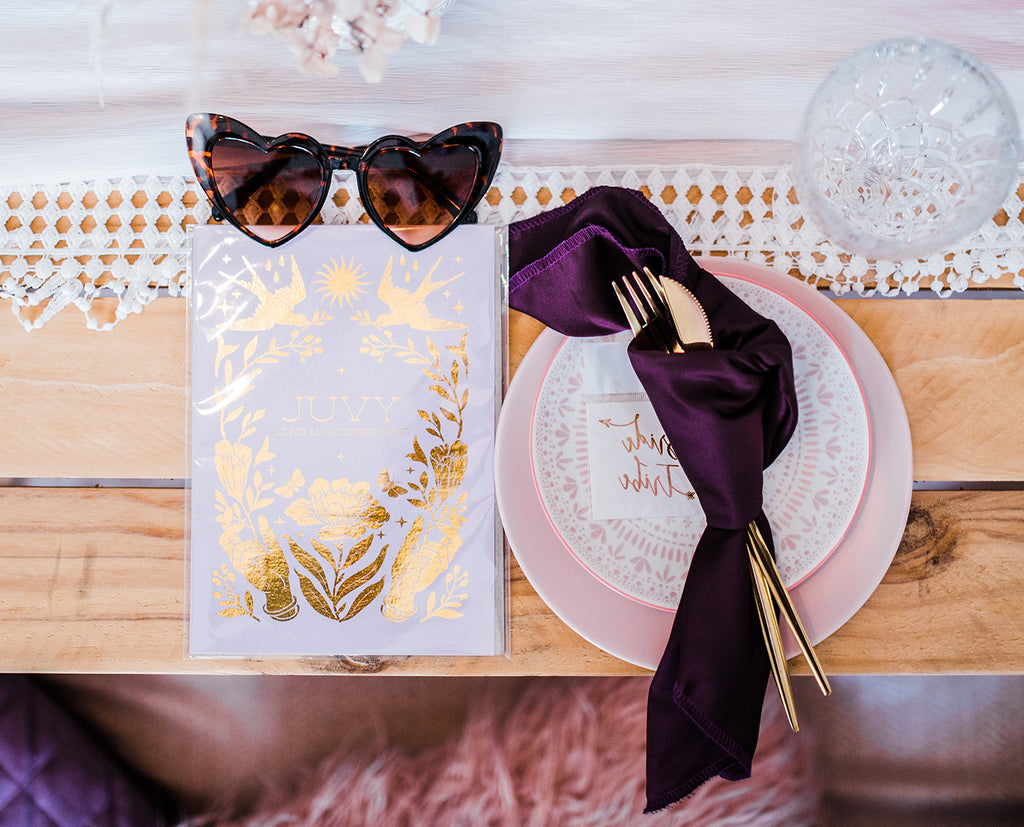 Dinner setting with placement holder of personalised gold foil print with hands, a flower, two doves and the sun. Sitting on top are heart shaped sunglasses. Gold cutlery is wrapped in a dark purple silk napkin