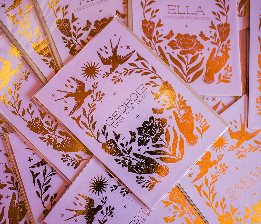 a scattered pile of personalised gold foil prints. Under each name, it says 'long live sisterhood' - the image on the print is of two open hands seperated by a flower, above the name are two doves holding vines with the sun in the middle