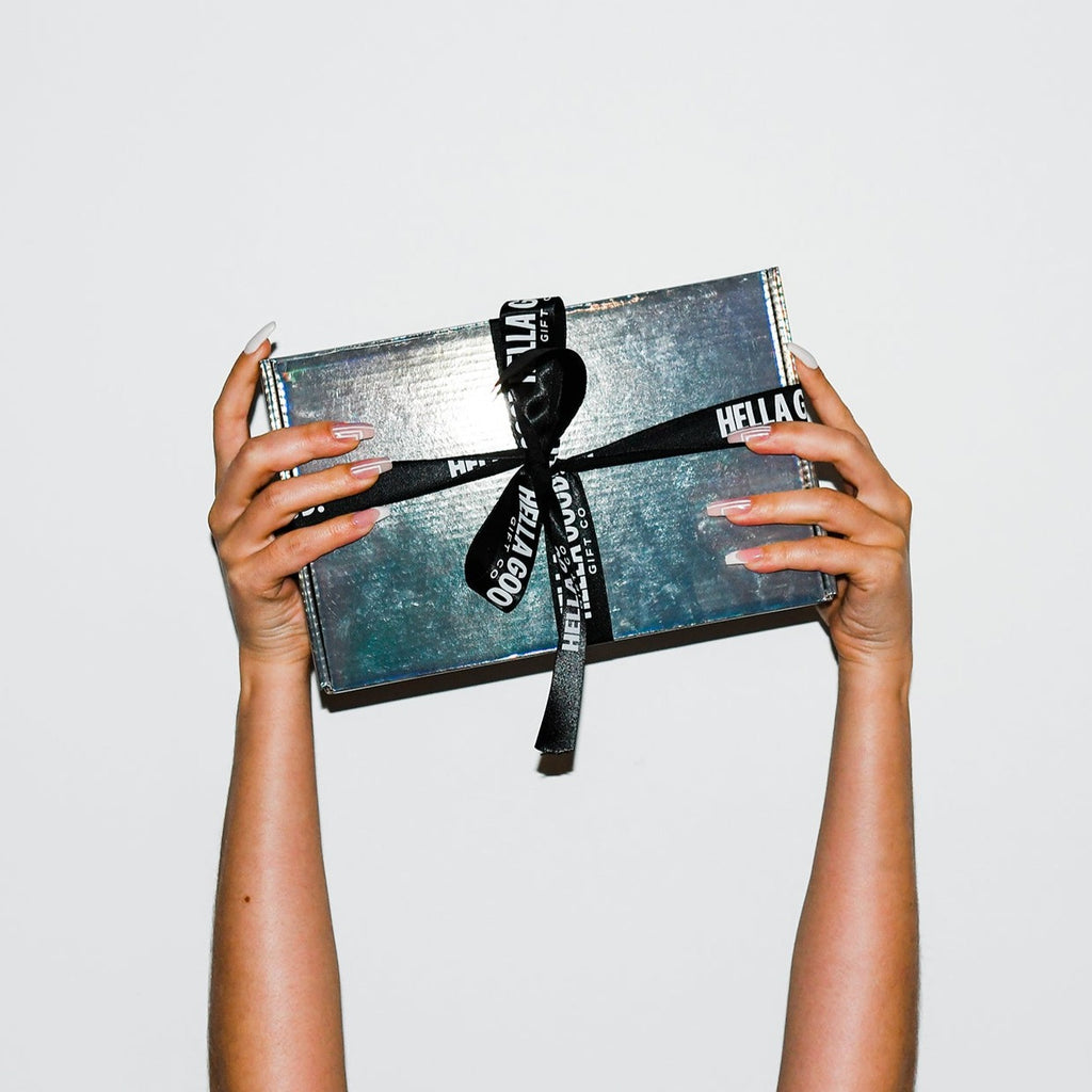two hands holding up a holographic box with hella good gift co ribbon wrapped around. 